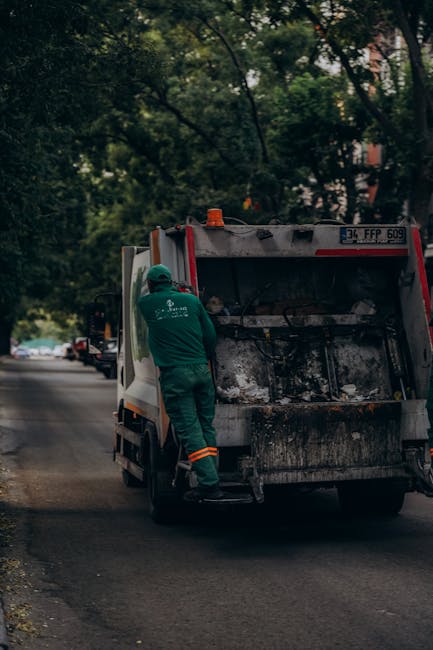 A sanitation worker wearing a green uniform and safety helmet is seen emptying a large black and white waste disposal dumpster positioned on a residential street. The dumpster is slightly dirty with some smudges and residue visible on its exterior. The worker is standing on the pavement next to the dumpster, holding a tool or handle to lift or operate the lid, which is open, revealing some waste materials inside. The street is lined with green trees providing partial shade and some parked cars are visible further down the road. The background includes a mix of leafy foliage and residential buildings, indicating a suburban setting. The scene captures a routine waste collection process typical of city or suburban waste management services, and it subtly relates to the logistics of waste and rubbish disposal that may be involved in house removals or relocations, especially when disposing of unwanted items during a move. Man with Van Mayfair occasionally manages such logistical tasks related to home relocation and moving services, supporting the safe transportation of belongings and waste clearance.