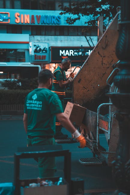 Two workers from Man with Van Mayfair are engaged in a home relocation process during dusk, loading cardboard boxes into a large, orange waste disposal vehicle parked on the street. The worker in the foreground, wearing a teal uniform with the company's logo and orange gloves, holds a box while standing next to a trolley filled with packing materials. The second worker, dressed in a dark green shirt with orange accents, is inside the vehicle, organizing boxes for transport. In the background, illuminated shop signs and digital billboards are visible, indicating a commercial area in Mayfair, London. The scene captures the detailed activity involved in furniture transport and packing during a house move, with the vehicle's open rear door revealing the loading process, and ambient street lighting providing clarity for the visually impaired. This image exemplifies the logistics of professional removals, highlighting box handling, secure packaging, and efficient loading within an urban moving context.