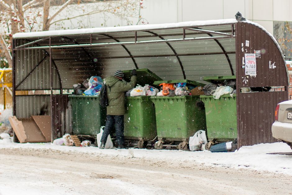 A sanitation worker wearing a green uniform and safety helmet is seen emptying a large black and white waste disposal dumpster positioned on a residential street. The dumpster is slightly dirty with some smudges and residue visible on its exterior. The worker is standing on the pavement next to the dumpster, holding a tool or handle to lift or operate the lid, which is open, revealing some waste materials inside. The street is lined with green trees providing partial shade and some parked cars are visible further down the road. The background includes a mix of leafy foliage and residential buildings, indicating a suburban setting. The scene captures a routine waste collection process typical of city or suburban waste management services, and it subtly relates to the logistics of waste and rubbish disposal that may be involved in house removals or relocations, especially when disposing of unwanted items during a move. Man with Van Mayfair occasionally manages such logistical tasks related to home relocation and moving services, supporting the safe transportation of belongings and waste clearance.