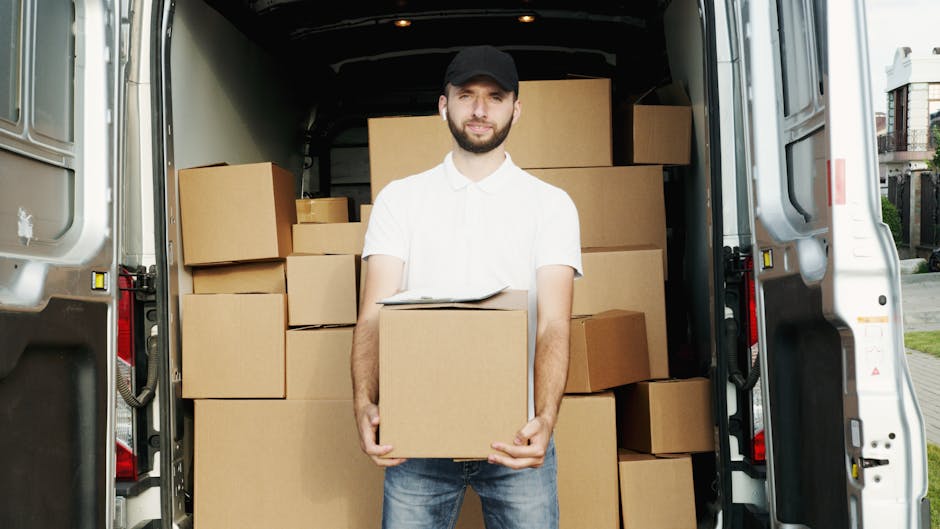 A person dressed in dark clothing, including a cap and sneakers, is standing at the open rear doors of a large white van, which is loaded with numerous cardboard boxes of varying sizes, some sealed wi