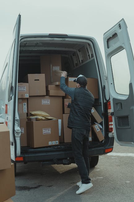 A man wearing a dark jacket, grey trousers, a black cap, and white sneakers is seen from behind during a home relocation, as he loads cardboard boxes into the open rear of a large white moving van parked on a paved outdoor area. The boxes vary in size and are sealed with packing tape, with some displaying labels or symbols indicating fragile contents. The van's interior is visible, revealing a spacious cargo area with additional boxes stacked towards the front. The man appears to be carefully placing a box onto the remaining contents inside the van, which is part of a professional moving process carried out by Man with Van Mayfair. The scene is well-lit with natural daylight, highlighting the careful packing and loading involved in furniture transport, packing, and moving logistics aimed at home relocation services.