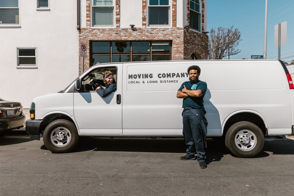 A male moving crew member stands in front of a white delivery van with the lettering 'MOVING COMPANY LOCAL & LONG DISTANCE' on its side, parked on a city street in front of a brick and white building with multiple windows. Inside the van's open driver-side window, a female colleague wearing gloves and sunglasses is visible, assisting with the loading process. The van is positioned close to the building's entrance, with some cardboard boxes and wrapped furniture items visible through the open rear doors of the vehicle. The scene occurs in daylight, with clear weather and a blue sky, reflecting an organized home relocation or furniture transport operation conducted by Man with Van Mayfair, a professional removals service provider specializing in packing and moving services.