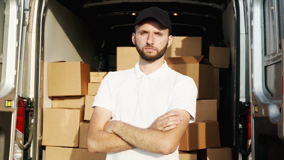 A young man with a serious expression, wearing a black cap and a white polo shirt, stands with his arms crossed inside the open rear of a large moving van. The van is filled with numerous cardboard boxes of various sizes, some wrapped in protective plastic or packing paper, indicating a home relocation process. The boxes are stacked neatly towards the back of the vehicle, which is parked near a building or on a street. The setting suggests the loading or unloading phase of furniture transport and packing during a professional house move in Mayfair, with the focus on the man representing the moving service, Man with Van Mayfair, involved in the moving logistics for a client relocating from Bond Street to Berkeley Square.