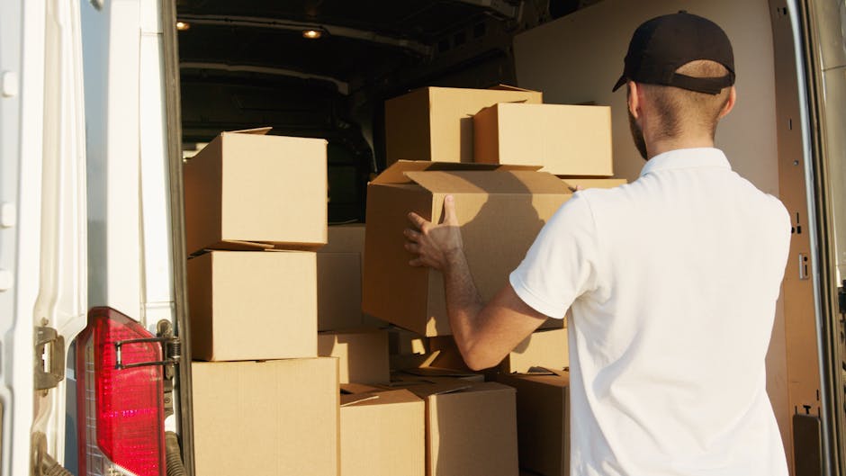 A young man with a serious expression, wearing a black cap and a white polo shirt, stands with his arms crossed inside the open rear of a large moving van. The van is filled with numerous cardboard boxes of various sizes, some wrapped in protective plastic or packing paper, indicating a home relocation process. The boxes are stacked neatly towards the back of the vehicle, which is parked near a building or on a street. The setting suggests the loading or unloading phase of furniture transport and packing during a professional house move in Mayfair, with the focus on the man representing the moving service, Man with Van Mayfair, involved in the moving logistics for a client relocating from Bond Street to Berkeley Square.
