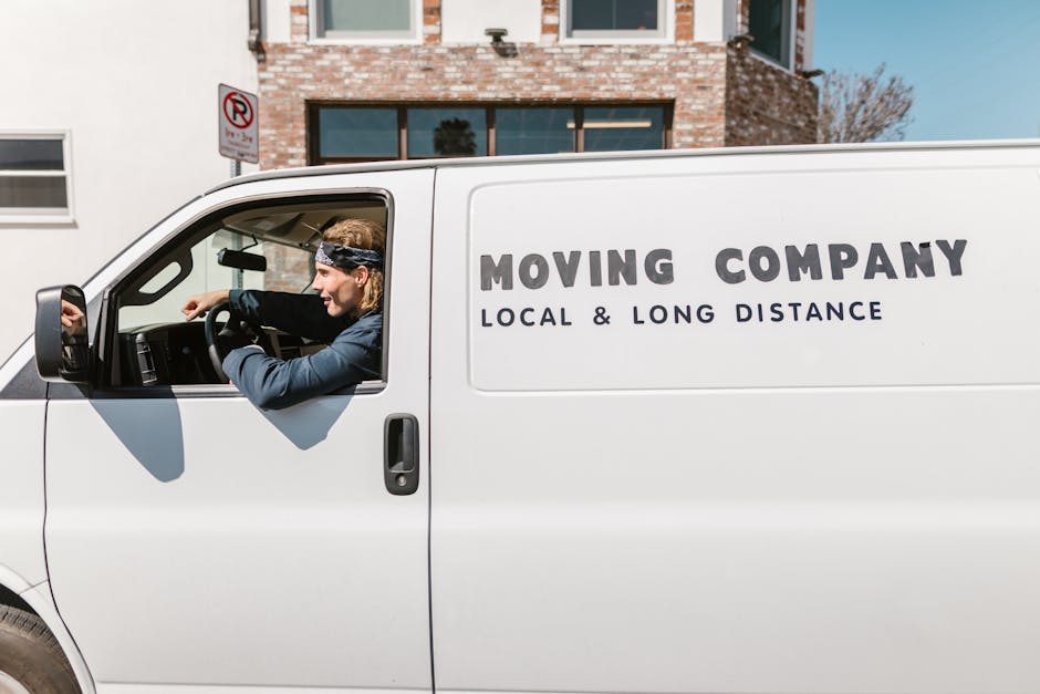 A white moving van parked on a residential street in Mayfair, with a man with long hair, wearing a bandana and casual clothing, sitting in the driver’s seat and preparing to load or unload items for a home relocation. The van has the words 'MOVING COMPANY' and 'LOCAL & LONG DISTANCE' printed on the side, indicating its service category. The vehicle is positioned near a modern brick building with large windows, and the street is clear with a no parking sign visible above. Inside the vehicle, various packing materials such as cardboard boxes, plastic wrap, and blankets can be seen on the floor and seats, ready for furniture transport. The environment is well-lit with natural daylight, capturing the logistics of packing and loading during the moving process, which is part of professional removal services provided by Man with Van Mayfair.