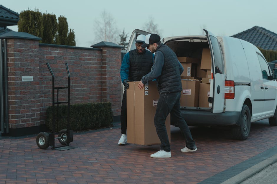 A white moving van parked on a residential street in Mayfair, with a man with long hair, wearing a bandana and casual clothing, sitting in the driver’s seat and preparing to load or unload items for a home relocation. The van has the words 'MOVING COMPANY' and 'LOCAL & LONG DISTANCE' printed on the side, indicating its service category. The vehicle is positioned near a modern brick building with large windows, and the street is clear with a no parking sign visible above. Inside the vehicle, various packing materials such as cardboard boxes, plastic wrap, and blankets can be seen on the floor and seats, ready for furniture transport. The environment is well-lit with natural daylight, capturing the logistics of packing and loading during the moving process, which is part of professional removal services provided by Man with Van Mayfair.