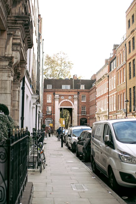 A city street scene in Mayfair featuring parked vehicles including a white van and several cars along the curb on the right side, with the van positioned closest to the foreground. The sidewalk on the left side displays bicycles secured to a black metal railing, with some boxes and packing materials visible nearby. In the background, a red brick building with an arched entryway and decorative white stone details frames the street, with a few people walking near the entrance and additional buildings extending further down the street. The environment suggests an urban area suitable for house removals or furniture transport, with the vans and packing supplies indicating an active home relocation process, supported by the professional services of Man with Van Mayfair.