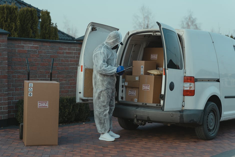 A person dressed in full protective PPE, including a white coverall, face mask, gloves, and shoe covers, standing beside an open white delivery van parked on a paved driveway next to a brick wall. Inside the van, there are several brown cardboard boxes of various sizes, some with fragile labels and handling symbols, neatly arranged on the van’s floor. One large box with a fragile sticker is placed on a small trolley outside the van, ready for carrying or loading. The person appears to be taking notes or inspecting the boxes, possibly as part of packing or inventory checks during a home relocation process. The background features trees and a slightly overcast sky, reflecting a typical suburban environment. This scene illustrates professional moving and packing services, highlighting the careful handling of belongings during a furniture transport or house removal, in line with the content of 'Packing and access tips for W1K flats in Mayfair' by Man with Van Mayfair.