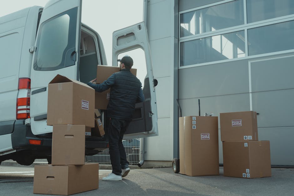 A man wearing a dark jacket and trousers is seen loading cardboard boxes into the rear of a white moving van parked outside a modern grey building with large glass windows. Several additional cardboard boxes, some labeled with fragile stickers, are placed on the ground nearby, with a small hand truck leaning against the building's exterior. The scene takes place during daylight, with natural lighting highlighting the presence of packing materials and transportation equipment used for house relocations. The man's activity is part of the furniture transport process associated with home relocation services provided by Man with Van Mayfair, as they handle packing and loading tasks for W1K flats in Mayfair. The image emphasizes the logistics involved in packing and access planning for efficient move management.