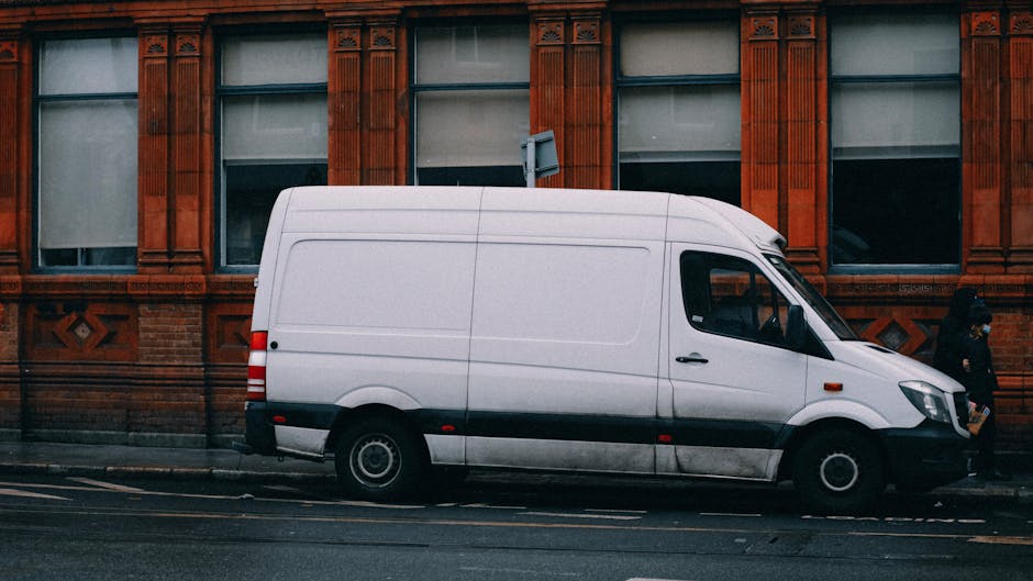 A white cargo van parked parallel to the pavement in front of a red-brick building with ornate window frames, part of a house removal or furniture transport operation. The van has tinted side windows and black detailing along the lower body. Inside the vehicle, various cardboard boxes and wrapping materials for packing are visible, indicating ongoing loading or unloading activities. A person dressed in dark clothing is walking past the van, possibly involved in the home relocation process, while another person is inside the van, arranging or carrying items. The surroundings include a dry pavement with lane markings, and the scene is illuminated with natural daylight, suggesting an urban street setting typical for professional removals near Grosvenor Square, Mayfair. The presence of equipment such as trolleys or straps is not visible in this image, but the setup aligns with vehicle-based furniture transport and external loading procedures supported by companies like Man with Van Mayfair.
