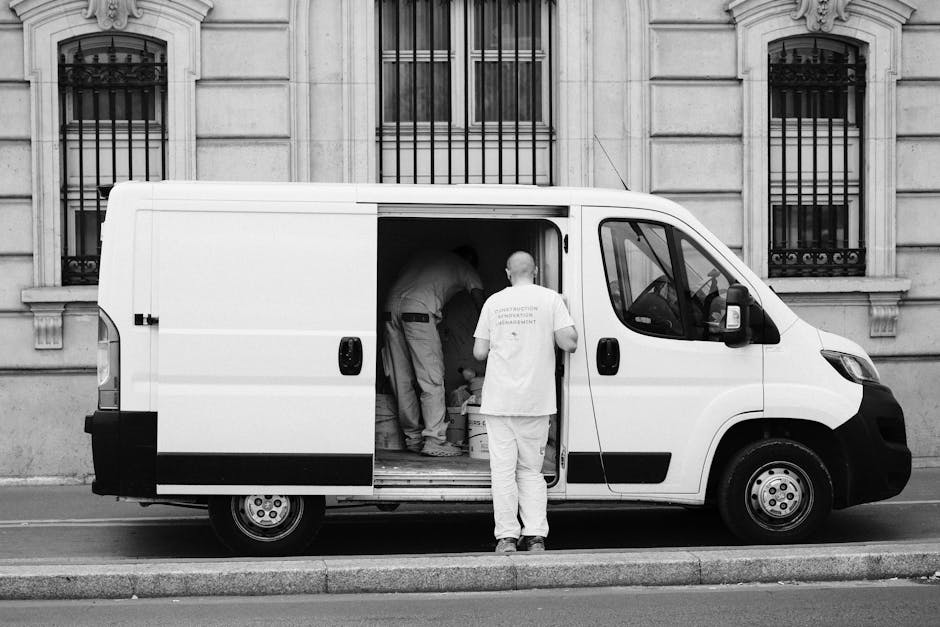 A man with short brown hair and a beard, dressed in a striped t-shirt and blue work overalls, is seated at the open side door of a white moving van. He is carefully examining a document or invoice while holding a cardboard box on his lap. Surrounding him on the pavement are several other cardboard boxes of various sizes, some stacked and some placed on the ground, which are likely used for packing belongings during a home relocation. The van, situated on a street near a curb with a concrete bollard visible in the foreground, has its sliding door fully open, revealing an interior suitable for furniture transport and packing materials. The scene captures a moment during the packing and loading process, typically associated with expert removals services such as those provided by Man with Van Mayfair, supporting the logistics of house removals near Grosvenor Square, Mayfair, with careful handling of packed items ready for transport to a new residence.