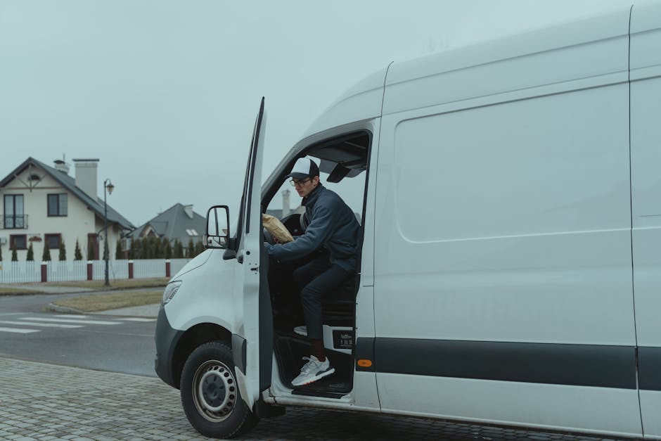 A man is seated inside the cab of a white delivery van, which is parked on a residential street. He is wearing a dark jacket, black trousers, and a black-and-white cap, and appears to be planning or reviewing a document while holding a brown paper package. The van's driver's side door is open, revealing the interior with some equipment visible, and the vehicle is positioned near the curb with the front wheel slightly turned toward the pavement. In the background, there are houses with pitched roofs, a white picket fence, and a streetlamp, all under a cloudy sky. The scene depicts the loading process typical of home relocation or furniture transport, with the van prepared for moving goods or household items, consistent with a professional removals service such as Man with Van Mayfair involved in same-day or last-minute moves.