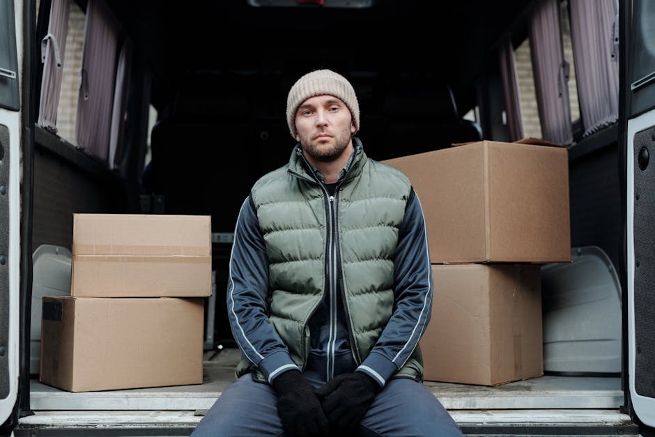 A young man with a neutral expression, wearing a beige knit beanie, a green puffer jacket with navy sleeves, dark gloves, and dark pants, is sitting on the edge of the open rear cargo area of a moving van. Inside the van, there are several cardboard boxes of various sizes stacked on the floor, some wrapped with plastic stretch film. The boxes are arranged in an orderly manner, with larger boxes placed at the bottom and smaller ones on top. The van's interior features dark panels, and the cargo area is partially visible with a glimpse of a ramp or floor at the back. The image depicts the process of home relocation, highlighting the loading or unloading stage of furniture transport and packing material handling, in natural daylight. Man with Van Mayfair specializes in removals and moving logistics, providing professional packing and transportation services, including loading processes with equipment like trolleys and blankets to protect furniture during transport.