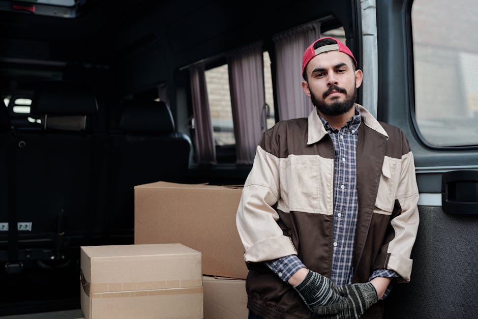 A young man with a neutral expression, wearing a beige knit beanie, a green puffer jacket with navy sleeves, dark gloves, and dark pants, is sitting on the edge of the open rear cargo area of a moving van. Inside the van, there are several cardboard boxes of various sizes stacked on the floor, some wrapped with plastic stretch film. The boxes are arranged in an orderly manner, with larger boxes placed at the bottom and smaller ones on top. The van's interior features dark panels, and the cargo area is partially visible with a glimpse of a ramp or floor at the back. The image depicts the process of home relocation, highlighting the loading or unloading stage of furniture transport and packing material handling, in natural daylight. Man with Van Mayfair specializes in removals and moving logistics, providing professional packing and transportation services, including loading processes with equipment like trolleys and blankets to protect furniture during transport.