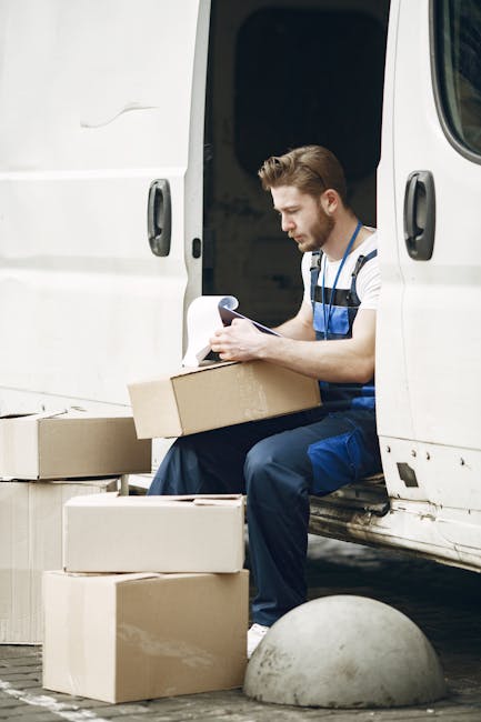 A male mover from Man with Van Mayfair is sitting on the edge of an open white van door during a home relocation process. He has a short beard and is wearing a striped t-shirt and blue overalls, holding a clipboard and pen while inspecting or organizing boxes. Several cardboard boxes of various sizes are around him, some stacked on the ground and others inside the van, which is parked on a brick pavement. The scene is set outdoors near a pavement with a concrete bollard visible in the foreground. The interior of the van appears dark, with the side door open, revealing part of the vehicle's interior. This image reflects the packing, loading, and logistical aspects of furniture transport and house removals, consistent with professional moving services provided by Man with Van Mayfair, particularly related to securing permits for residential moving in Westminster.