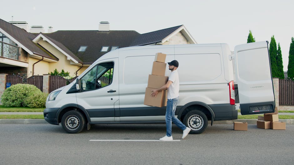 A person wearing a white t-shirt, jeans, and a black cap is engaged in home relocation activities by loading cardboard boxes into the back of a white panel van parked on a residential street. The individual is carrying a stack of three cardboard boxes, with additional boxes placed on the ground next to the van’s open rear door. The van is positioned parallel to a sidewalk, with a modern suburban house featuring a pitched roof, skylights, and a fenced garden visible in the background. The scene is set during daylight hours, with natural lighting clearly illuminating the moving process. Man with Van Mayfair, a professional removals company, is involved in furniture transport and packing and moving services, demonstrating the logistics of a house removal or relocation process.